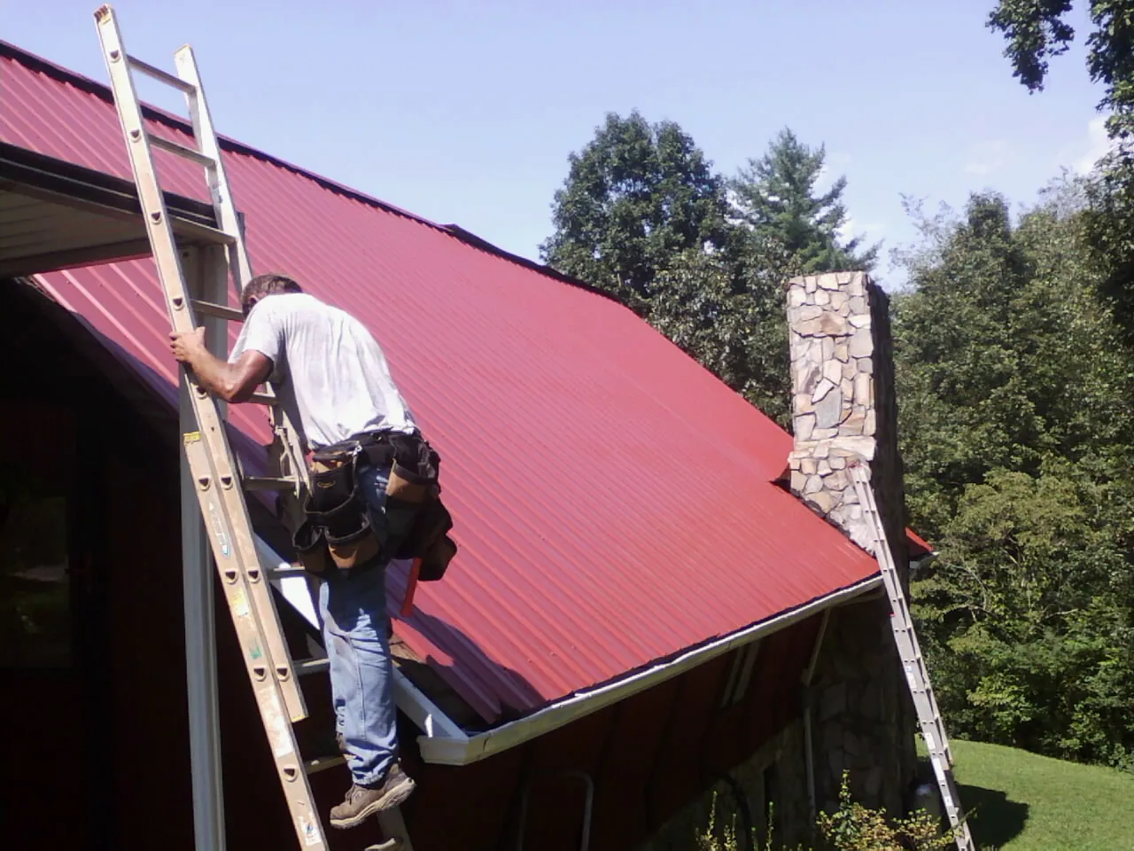 Roofer on a ladder beside a red metal roof and stone chimney — Soffit & Fascia Repair by Roofing Brothers in Stantonville